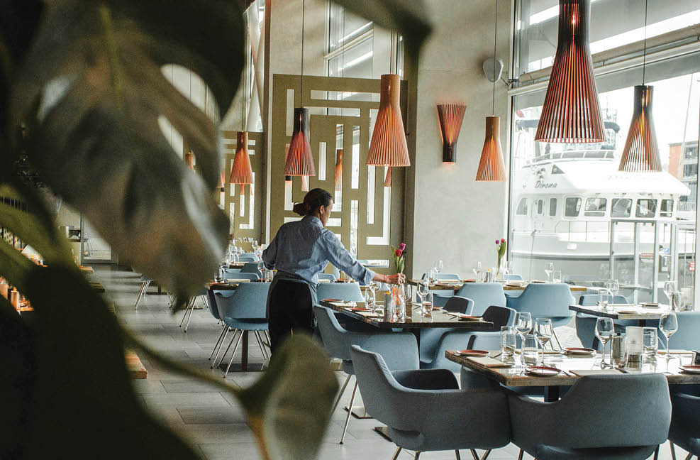 A man is standing in a restaurant, wearing a blue shirt. The restaurant has a large window and several chairs and dining tables. There are also potted plants and vases in the room. AI generated content