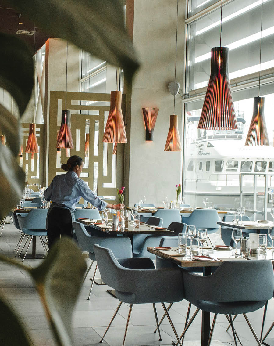 A man is standing in a restaurant, wearing a blue shirt. The restaurant has a large window and several chairs and dining tables. There are also potted plants and vases in the room. AI generated content
